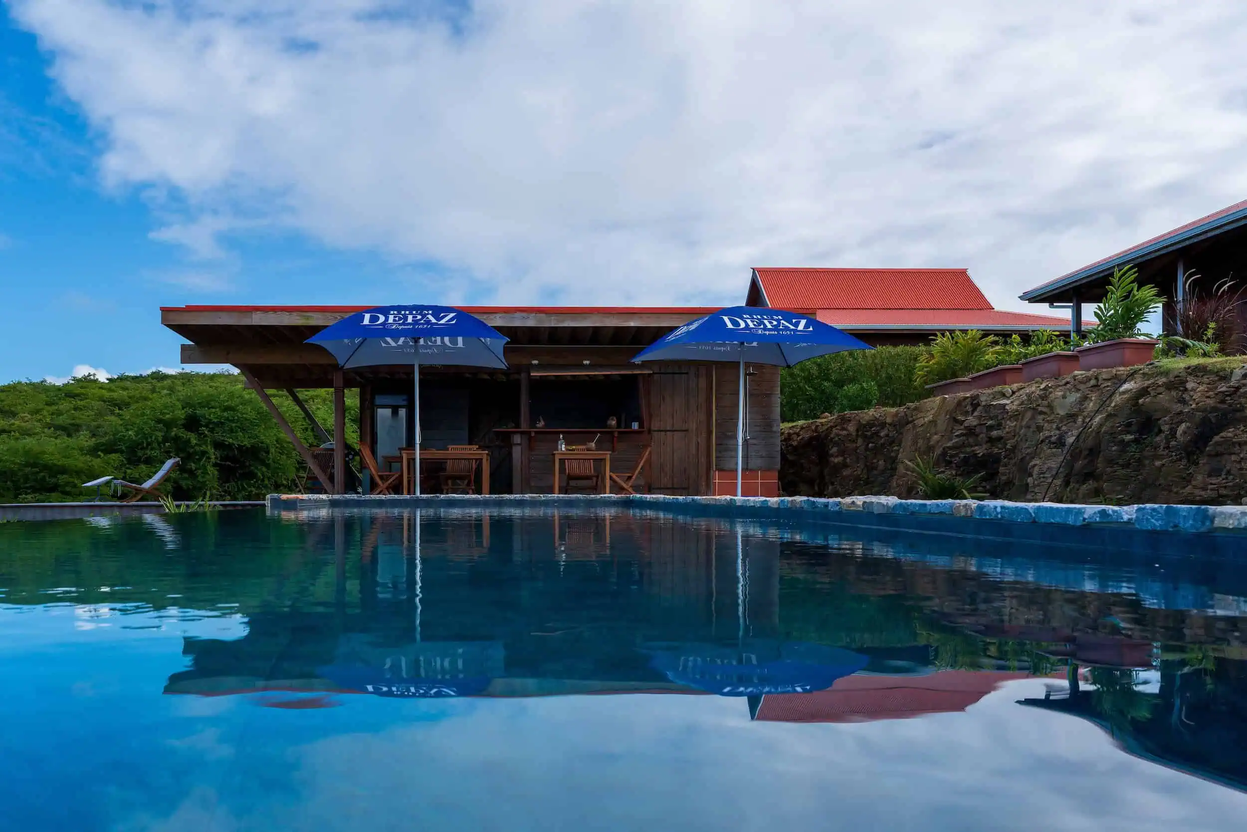 piscine avec en arrière plan le carbet avec un bar et des tables avec des parasols depaz 