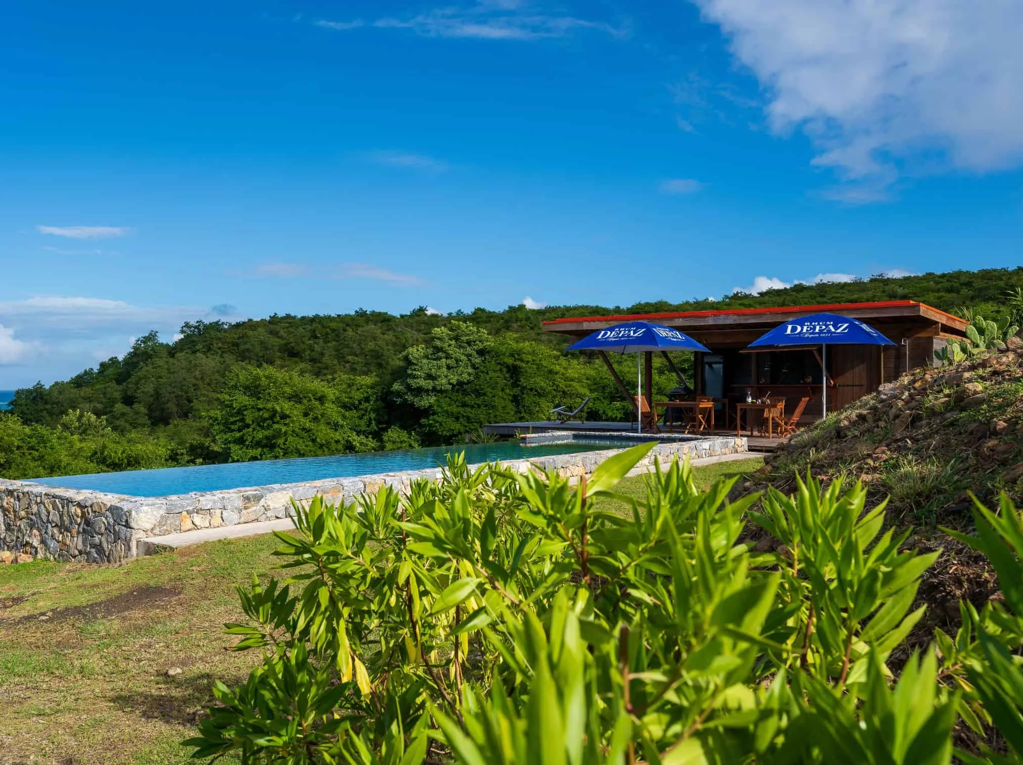 piscine_a_debordement_entouree_de_verdure_avec_une_cabane_en_bois_et_des_parasols_bleus_marques_depaz piscine_a_debordement_entouree_de_verdure_avec_une_cabane_en_bois_et_des_parasols_bleus_marques_depaz dans nos Écolodges d’exception au cœur de la Martinique