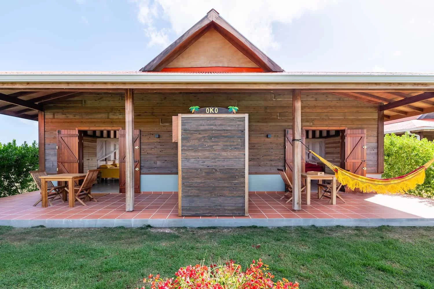 Maison en bois avec deux portes ouvertes, terrasse carrelée avec mobilier en bois et un hamac jaune suspendu, jardin fleuri au premier plan.